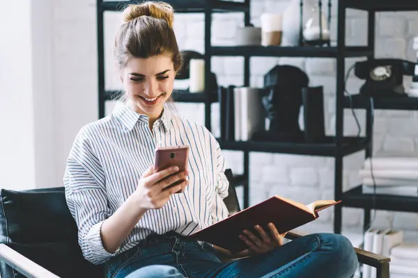 Cheerful young woman laughing while reading received message on smartphone sitting with textbook on chair in flat with stylish home interior.Positive female watching funny video in blog on cellular
