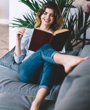 Evde eğlenceli vakit geçiren neşeli genç bir kadın. Eğlenceli bir kitapla eğleniyor. Neşeli hippi kız rahat bir kanepede uzanıyor ve modern dairede ders kitabı okuyor.