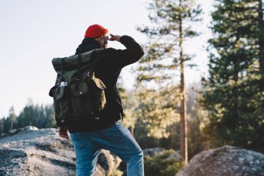 Back view of male traveler with backpack standing on peak of mountain getting to destination in tour, guy wanderlust exploring wild nature and admire breathtaking scenery of landscape from rock clipart