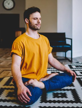Meditasyon sırasında gülümseyen genç bir adam modern apartmanda rahat bir halının üzerinde poz veriyor. 20 yaşında, yoga yapan sakin bir hippi.