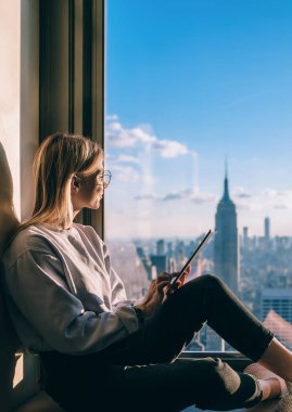 Gülümseyen hippi kız, New York şehrinin güzel manzarasının keyfini çıkarırken aynı zamanda gökdelenlerin en üst katındaki otel odasında oturuyor. Tatil boyunca iletişim için dokunmatik ped kullanan mutlu kadın turist.