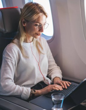 Beyaz bir kadın uçuş sırasında serbest çalışıyor ve kablosuz internet erişimini kullanıyor. Genç sarışın kadın müzik dinliyor, klavye e e-postası uçaktan iniyor.