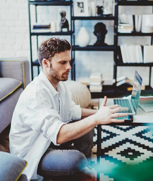 Young man dressed in casual wear sitting on floor and typing text message on smartphone device while waiting for online conference with colleagues, millennial male freelancer connected to 4g wireless