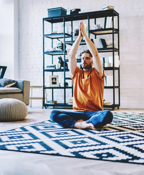 Calm young bearded man sitting in lotus pose on cozy carpet and holding hands in namaste above head during meditation at home apartment.Hipster guy with closed eyes doing yoga exercises