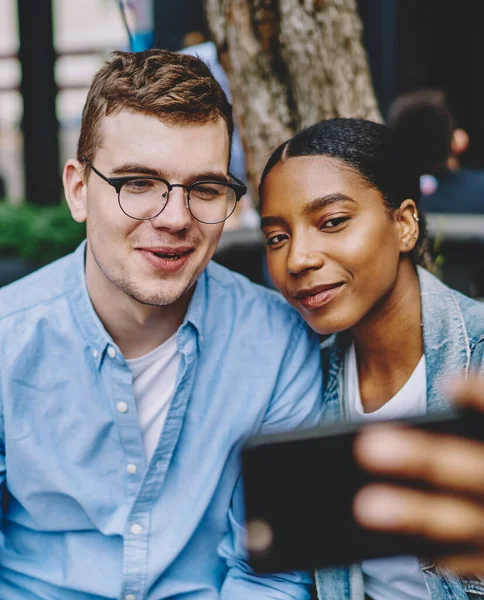 Happy multicultural young marriage smiling at front camera of modern smartphone and making selfie for social networks sitting in urban setting.Diverse friends taking photo on mobile phone on street