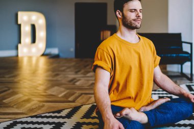 Meditasyon sırasında gülümseyen genç bir adam modern apartmanda rahat bir halının üzerinde poz veriyor. 20 yaşında, yoga yapan sakin bir hippi.