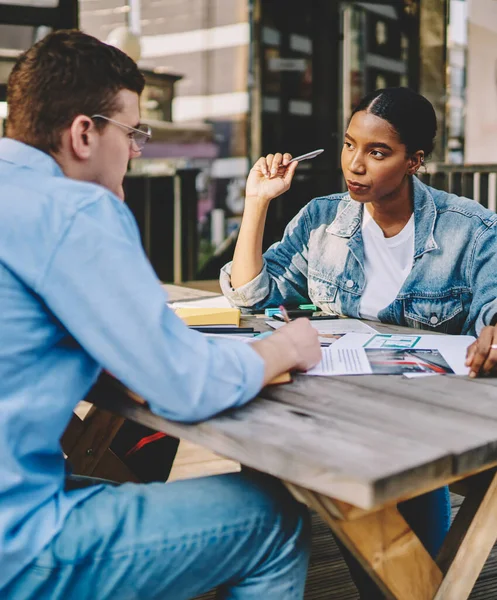 Young multiracial male and female colleagues having conversation about common project at cafe outdoors, confident businesswoman having meeting with man partner for finance consultancy of project
