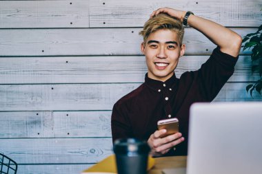 Mutlu Asyalı hippi öğrenci yüzünde sevimli bir gülümsemeyle kafeteryada dinlenirken elinde akıllı bir telefonla internete bağlı bir dizüstü bilgisayarla uzaktan çalışmaktan dolayı dinleniyor.