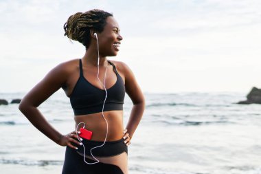 Aktif giyim giymiş sportif siyah kadın koşucu sabah seacoast ayakta telefona bağlı kulaklık müzik dinleme, koşmak için eşofman giymek dreads ile neşeli kadın