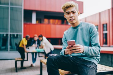 Sıradan giyinmiş Çinli bir gencin portresi kameraya bakarken açık havada oturan ve 4G internet bağlantısı kullanan modern akıllı telefon cihazına mesaj yazıyor. Japon hipster blogcu