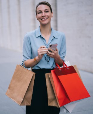 Heyecanlı kadın alıcının yarım boy portresi kağıt paketlerle tanıtım alanında duruyor ve kameraya gülümsüyor modaya uygun giyinmiş mutlu hippi kız elinde modern cep telefonu cihazıyla dışarıda bekliyor.