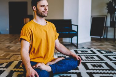 Meditasyon sırasında gülümseyen genç bir adam modern apartmanda rahat bir halının üzerinde poz veriyor. 20 yaşında, yoga yapan sakin bir hippi.