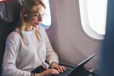 Beyaz bir kadın uçuş sırasında serbest çalışıyor ve kablosuz internet erişimini kullanıyor. Genç sarışın kadın müzik dinliyor, klavye e e-postası uçaktan iniyor.