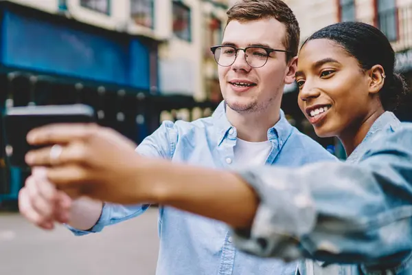 Happy multicultural young marriage making selfie picture on front camera of smartphone standing in urban setting.Positive dark skinned hipster girl with caucasian friend making photo on phone for blog