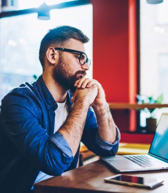 Ciddi bir erkek moda göz giyimi uzmanı içerik web sitesinde modern teknolojinin tebligatı hakkında haberleri okurken aynı zamanda kafenin içinde uzaktan çalışıyor ve ücretsiz internet bağlantısı kullanıyor.