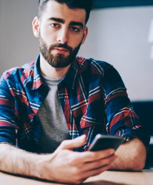 Ortak çalışma alanında masada oturan hücresel cihazsms mesajı gönderirken kameraya bakan sakallı genç adamın portresi. 4g internet kullanarak akıllı telefon aparatı hipster blogger yükleme