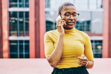 Gözlüklü Afrikalı Amerikalı kadın modern akıllı telefondaki telefon görüşmeleri sırasında şehir ortamında dolaşım bağlantısını kullanarak bazı bilgileri açıklıyor. Koyu tenli kadın cep telefonundan arıyor.