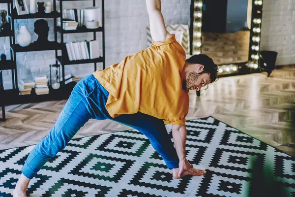 Young man doing sportive exercises during stretching training on carpet at home interior.Motivated hipster guy leads healthy lifestyle and maintains a vital tone in modern apartment