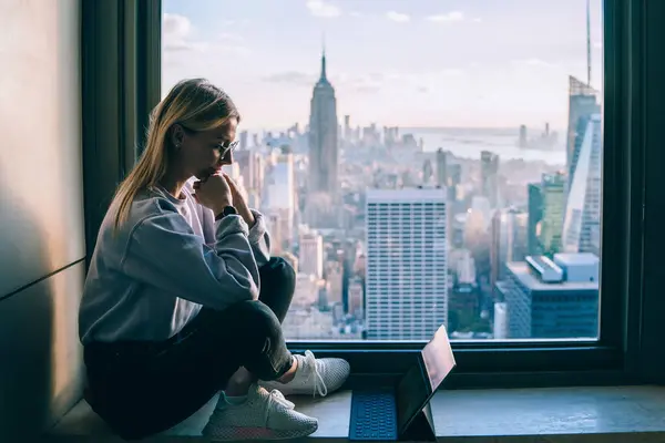 Pensive female office employee thinking on creating new project while sitting on window sill with scenery view of New York downtown. Hipster girl digital nomad working on modern tablet during travel
