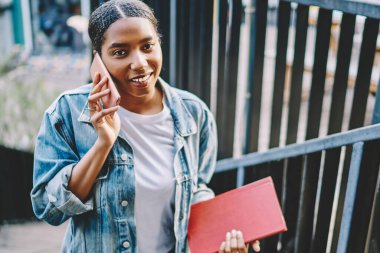 Akıllı telefondan ararken kameraya gülümseyen ve dışarıda cep telefonu operatörüyle konuşan güzel bir genç kadının portresi. Modern cep telefonuyla iletişim kuran güzel bir hipster blogcusu.