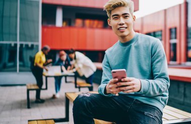 Açık havada oturan modern akıllı telefon cihazındaki uygulamayı güncellerken kameraya gülümseyen neşeli hipster blog yazarının portresi. Hücresel ağlarda sosyal ağlarda olumlu Japon genç adam sohbet ediyor.