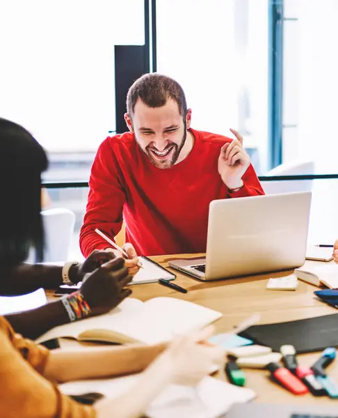 Cheerful male team leader laughing at meeting with students satisfied with friendly communication, excited joyful crew of employees having fun during workflow in coworking office sitting at table