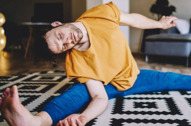 Gözleri kapalı genç bir adam halının üzerinde oturmuş, esneme egzersizleri yapıyor ve evde esneklik kazanıyor. Yoganın motive olmuş erkek aşığı, apartman dairesinde sabah antrenmanı yapıyor.