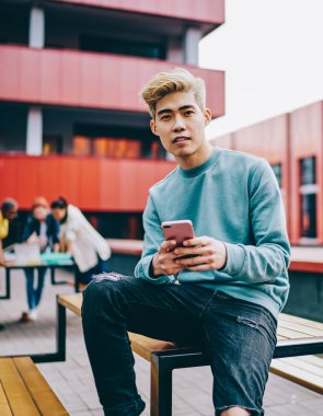 Sıradan giyinmiş Çinli bir gencin portresi kameraya bakarken açık havada oturan ve 4G internet bağlantısı kullanan modern akıllı telefon cihazına mesaj yazıyor. Japon hipster blogcu