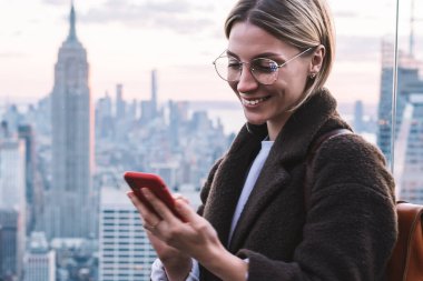 Gülümseyen beyaz kadın tatiller sırasında Empire State manzaralı binayla gözlem güvertesinde dikilirken cep telefonuyla uygulama kullanıyordu. New York 'ta mutlu kadın turist.