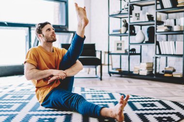 Genç adam bacak esnetiyor ve yukarı bakıyor. Modern apartman dairesinde spor egzersizleri yapıyor. Hipster adam esnek ve rahat halı üzerinde yoga yapıyor.