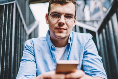 Merdiven başında oturan modern akıllı telefon cihazında 4G internet bağlantısını kullanırken kameraya bakan gözlüklü yakışıklı genç adamın portresi. Hipster blogcusu hücresel uygulamada profilini güncelliyor.
