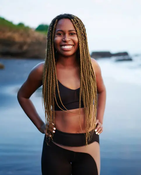 Okyanus suyu ile yıkanmış plajda ayakta dreads ile pozitif neşeli sportwoman Portresi, sahilde kamera gülümseyerek egzersiz için aktif giysiler giymiş mutlu koyu tenli genç kadın