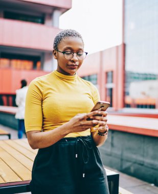 Gözlüklü Afro-Amerikan kadın elinde modern akıllı telefon ve cihazla şehir ortamında 4G internet bağlantısı kuruyor. Koyu tenli kadın cep telefonundan SMS mesajı alıyor.