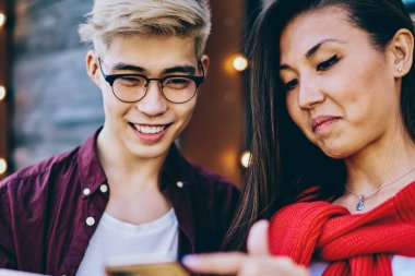 Neşeli hippi Asyalı arkadaşlar ellerinde modern akıllı telefonlar ve çevrimiçi video izliyorlar şehir ortamında dikiliyorlar, pozitif Japon çift hücresel ağlarda sosyal ağlarda tasarım profillerini tartışıyorlar.