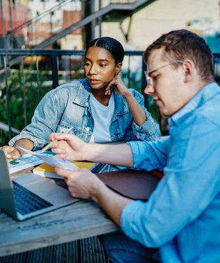 Yoğunlaşmış koyu tenli hippi öğrenci, modern dizüstü bilgisayarda beyaz meslektaşlarıyla birlikte ders izliyor. Tahta masada oturuyorlar ve seminer için hazırlanıyorlar.