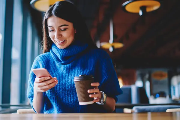 Carefree female hipster girl chatting with friends via modern mobile phone and holding cup with coffee, successful blogger installing application for communication and dialing number on mobile phone