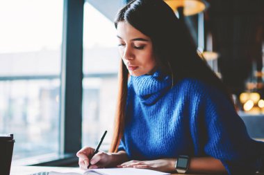 Ciddi düşünceli esmer hipster kız kafeteryada istirahat sırasında sınava hazırlanıyor, modern cep telefonu ahşap masada kalırken üniversite kampüsünde kız öğrenci öğrenme düşünme