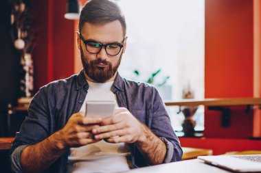 Görme düzeltmesi için gözlük takan ciddi bir erkek blogcu akıllı telefon aygıtında SMS 'leri okurken aynı zamanda laptoplu bilgisayarla birlikte kafenin iç kısmında vakit geçiriyor.