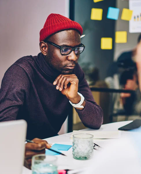 Serious african american hipster guy in spectacles concentrated on reading information during lesson, cropped image of pondering male employee analyzing accounting of company income on meeting
