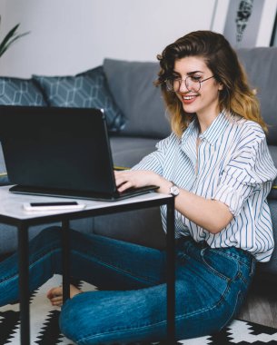 Gözlüklü neşeli genç bir kadın modern dizüstü bilgisayarda uzaktan çalışırken gülüyor. Ev içinde yerde oturuyor. İnternette arkadaşlarıyla sohbet eden pozitif hipster kız.