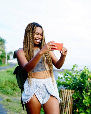 Sırt çantası akıllı telefon cihazüzerinde tropikal yerellik fotoğrafları yapma ile Neşeli afro Amerikan gezgin, yaz gezisi sırasında cep telefonu fotoğraf çekmekten dreads ile pozitif koyu tenli turist