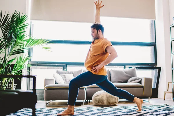 Side view of sportive young man holding hand up during morning workout at home apartment.Hipster guy warming up before yoga training on stylish carpet in flat enjoying harmony and healthy vitality