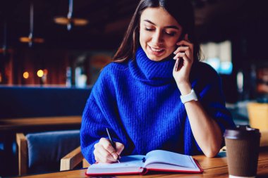 Kampüs kafede okurken arkadaşına cep telefonu yla arayan neşeli kadın, 4g internete bağlı gülümseyen hipster kız notlar yazarken telefonla konuşuyor ve kafede oturuyor