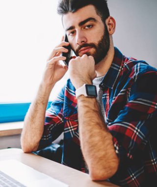 Mesafe iş için dizüstü bilgisayar ile masaüstünde oturan kamera bakarken akıllı telefon arama sakallı hipster öğrenci portresi. Genç adam serbest netbook de hücresel çalışma konuşuyor