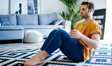 20 yaşında genç bir adam, halıda oturuyor ve modern apartmanlarda sağlıklı vücut ve canlılık için sabah egzersizleri yapıyor. Motive olmuş Kafkasyalı hipster çocuk evde fiziksel antrenman yapıyor.