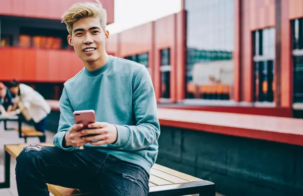 Açık havada oturan modern akıllı telefon cihazındaki uygulamayı güncellerken kameraya gülümseyen neşeli hipster blog yazarının portresi. Hücresel ağlarda sosyal ağlarda olumlu Japon genç adam sohbet ediyor.