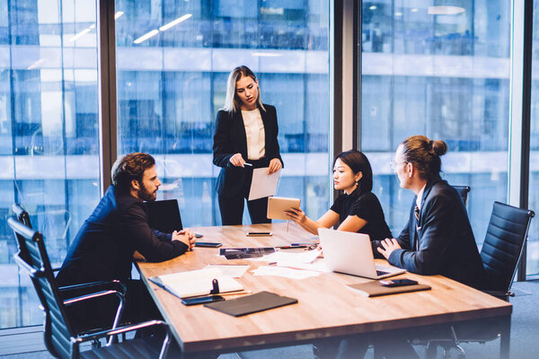 Предпринимательница, объясняющая стратегию компании профессиональной команде сотрудников. Женщина-предприниматель представляет финансовый отчет корпоративным коллегам, стоящим возле стола заседаний в офисном интерьере