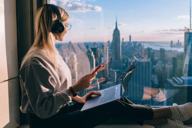 Dizüstü bilgisayardaki serbest çalışma sırasında kulaklıkla müzik dinleyen sarışın kadın blogcu. Hippi kız gezgin New York şehir merkezinin panoramik manzarasının keyfini çıkarıyor ve sesli kitapla çalışıyor.