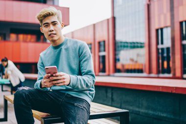 Sıradan giyinmiş Çinli bir gencin portresi kameraya bakarken açık havada oturan ve 4G internet bağlantısı kullanan modern akıllı telefon cihazına mesaj yazıyor. Japon hipster blogcu