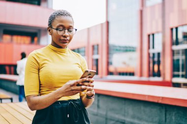 Gözlüklü Afro-Amerikan kadın elinde modern akıllı telefon ve cihazla şehir ortamında 4G internet bağlantısı kuruyor. Koyu tenli kadın cep telefonundan SMS mesajı alıyor.